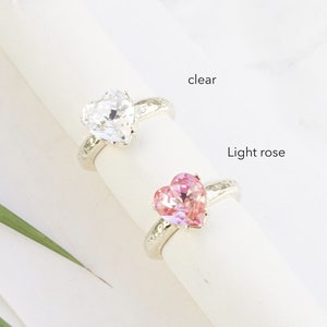 May include: Two rings with heart-shaped stones on a white display. One ring features a clear stone, and the other features a light rose stone. Both rings have a textured, shiny silver band. The text labels clear, Light rose, and Shiny silver appear.