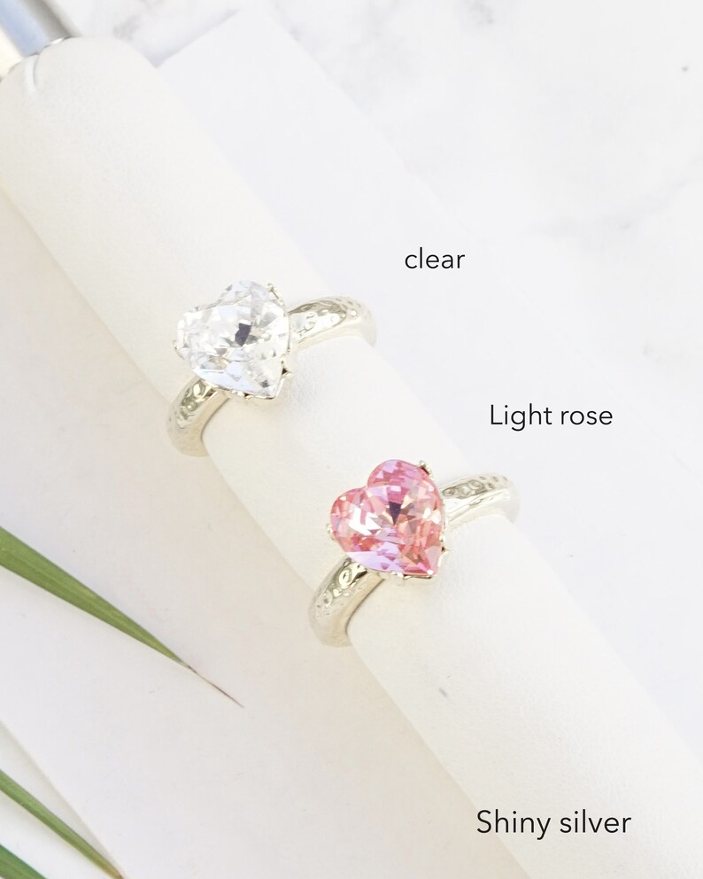May include: Two rings with heart-shaped stones on a white display. One ring features a clear stone, and the other features a light rose stone. Both rings have a textured, shiny silver band. The text labels clear, Light rose, and Shiny silver appear.