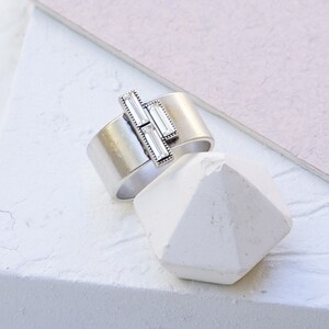 Puede incluir: Un anillo de plata con tres piedras de cristal transparente engastadas en un dise&ntilde;o geom&eacute;trico. El anillo est&aacute; sobre un fondo blanco.