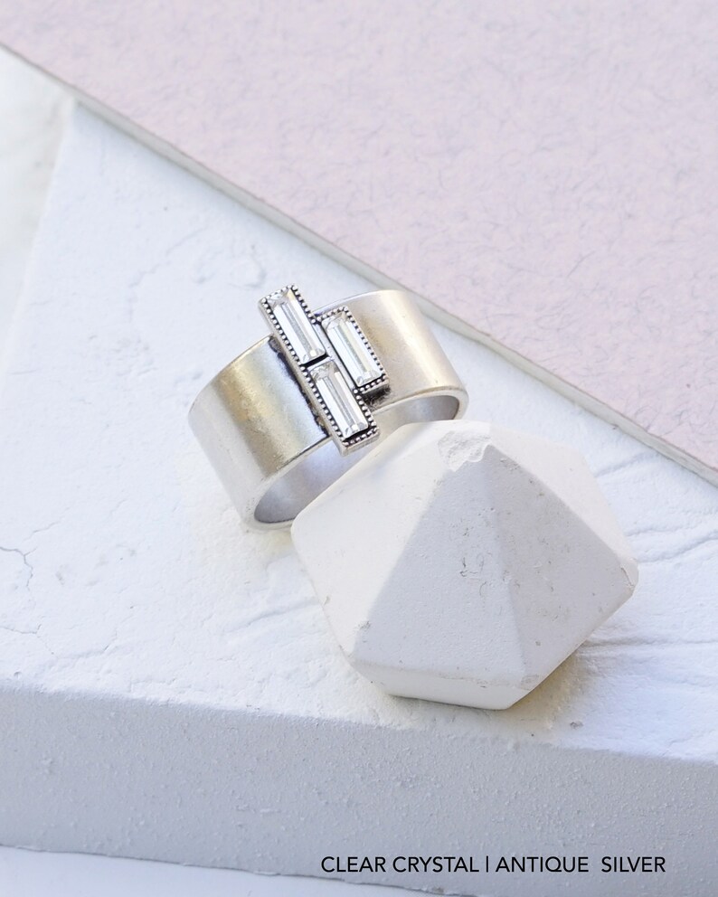 Puede incluir: Un anillo de plata con tres piedras de cristal transparente engastadas en un dise&ntilde;o geom&eacute;trico. El anillo est&aacute; sobre un fondo blanco.