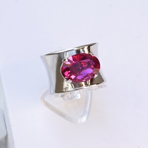 May include: A wide silver-toned ring featuring a large, oval-cut fuchsia gemstone set in the centre. The band has a smooth, reflective metallic finish. The text "Fuchsia with Shiny silver finish" is printed at the bottom of the image.