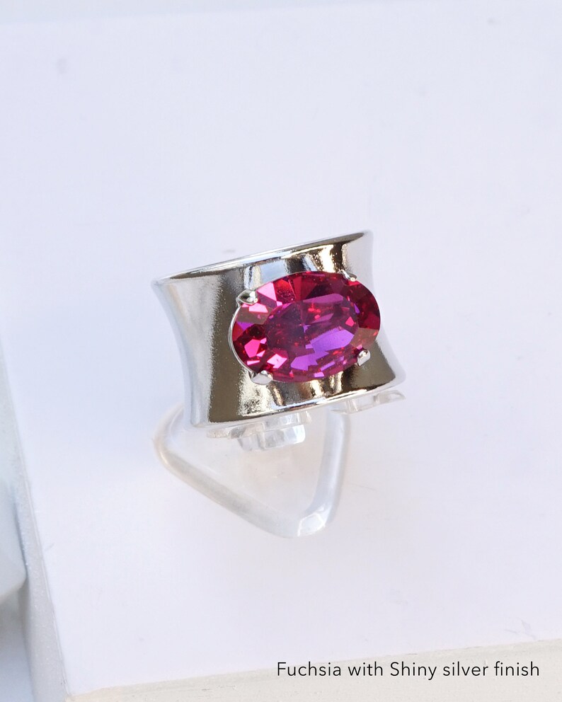 May include: A wide silver-toned ring featuring a large, oval-cut fuchsia gemstone set in the centre. The band has a smooth, reflective metallic finish. The text "Fuchsia with Shiny silver finish" is printed at the bottom of the image.