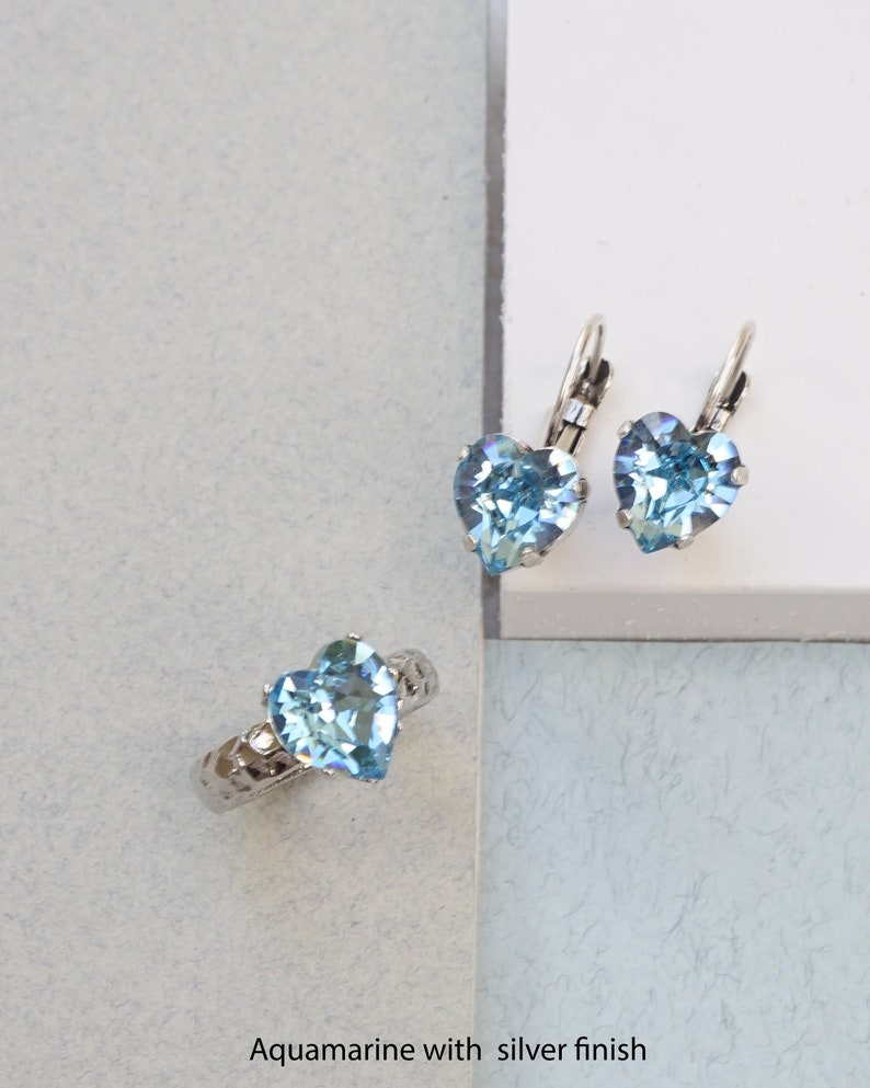 May include: A silver-toned ring and a pair of leverback earrings, each featuring a faceted heart-shaped aquamarine-colored stone. The ring band has a textured, hammered finish. The text "Aquamarine with silver finish" is printed at the bottom.