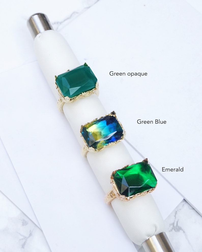 K&ouml;nnte beinhalten: Drei goldene Ringe mit verschiedenen farbigen Steinen. Der obere Ring hat einen undurchsichtigen gr&uuml;nen Stein, der mittlere Ring hat einen gr&uuml;n-blauen Stein und der untere Ring hat einen dunkelgr&uuml;nen Stein. Die Ringe sind mit "Gr&uuml;n undurchsichtig", "Gr&uuml;n Blau" und "Smaragd" beschriftet.