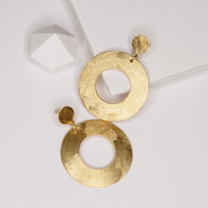 May include: A pair of matte gold hoop earrings with a textured finish. The earrings are circular with a smaller circle cut out in the center.