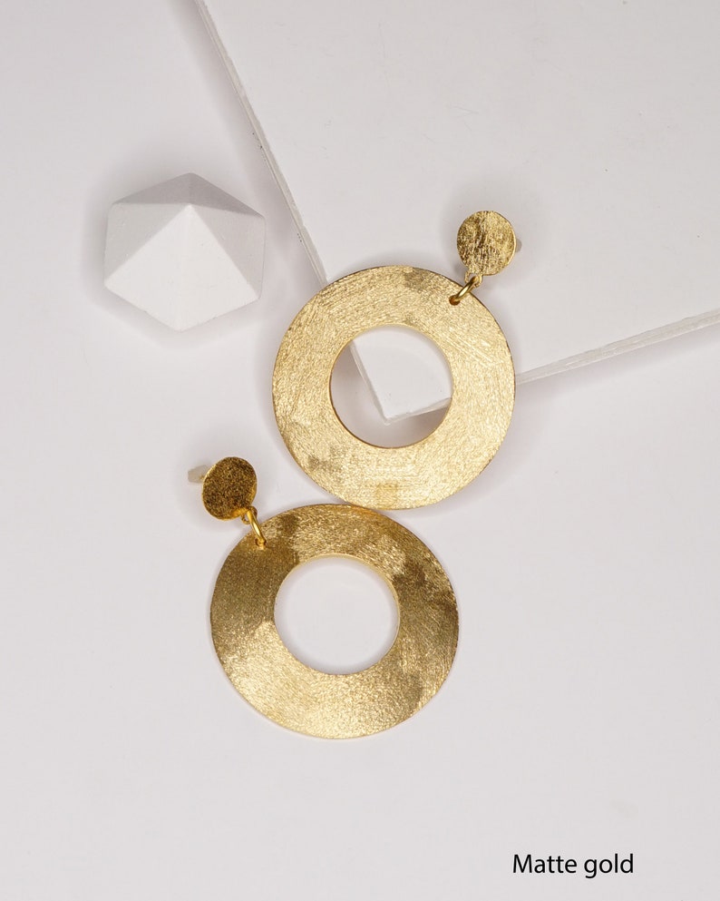 May include: A pair of matte gold hoop earrings with a textured finish. The earrings are circular with a smaller circle cut out in the center.