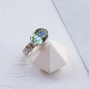 May include: A ring featuring an oval-shaped Erinite green crystal set in an antique silver band with a hammered, textured finish. The ring is displayed resting against a white geometric object. The text Erinite | Antique silver is visible.