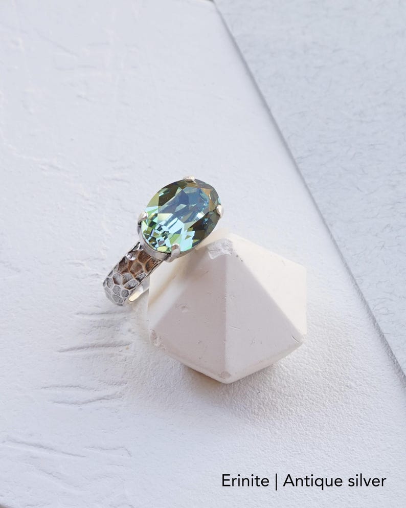 May include: A ring featuring an oval-shaped Erinite green crystal set in an antique silver band with a hammered, textured finish. The ring is displayed resting against a white geometric object. The text Erinite | Antique silver is visible.
