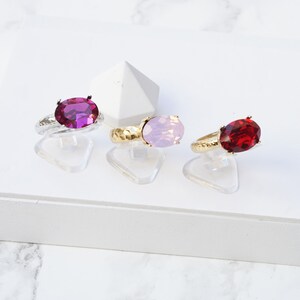 May include: Three rings with oval-cut faceted stones in fuchsia, light pink opal, and deep red light siam colors. Each ring features a textured metallic band and is displayed on a clear stand. Text labels below identify the colors as Fuchsia, Pink opal, and Light Siam.