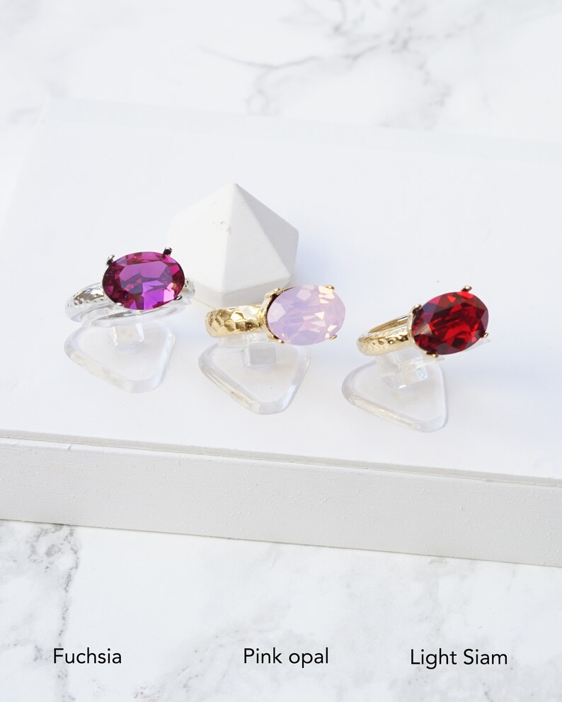 May include: Three rings with oval-cut faceted stones in fuchsia, light pink opal, and deep red light siam colors. Each ring features a textured metallic band and is displayed on a clear stand. Text labels below identify the colors as Fuchsia, Pink opal, and Light Siam.