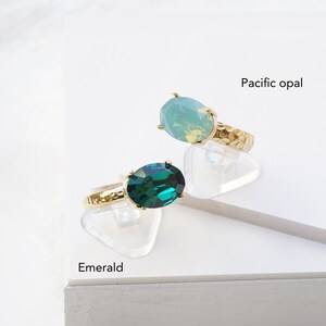 May include: Two gold-toned rings with hammered bands, each featuring a large oval-cut stone. One ring has a deep green stone labeled Emerald, and the other has a light blue-green stone labeled Pacific opal. Both rings are set on clear stands.