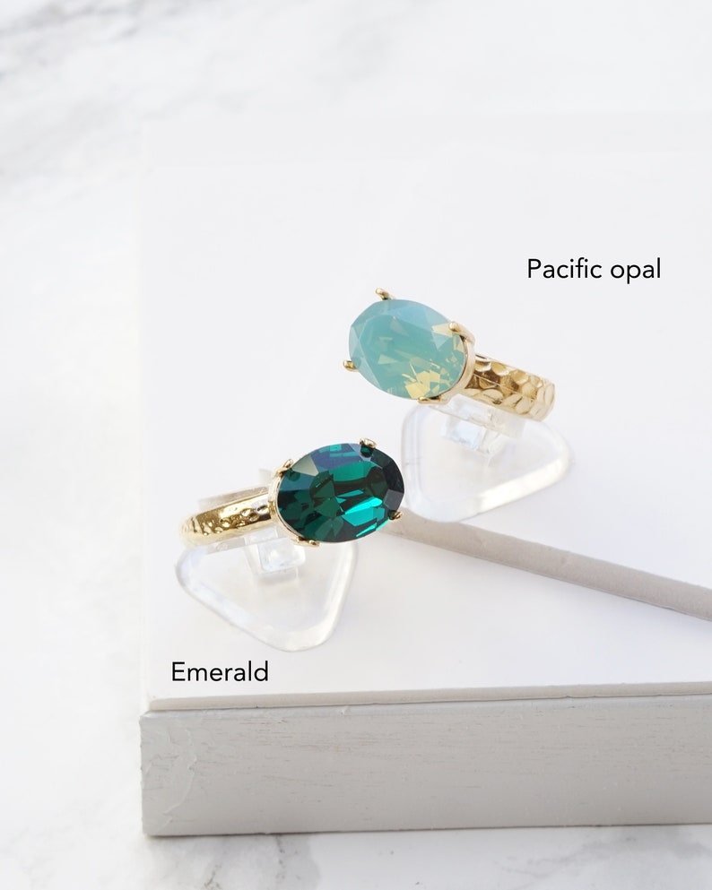 May include: Two gold-toned rings with hammered bands, each featuring a large oval-cut stone. One ring has a deep green stone labeled Emerald, and the other has a light blue-green stone labeled Pacific opal. Both rings are set on clear stands.