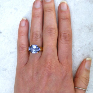 May include: A gold ring featuring a large, oval-cut, light purple gemstone set in a prong setting on the middle finger, accompanied by a thin, plain silver-toned band worn on the thumb, showcasing a contrast between the two distinct ring styles.