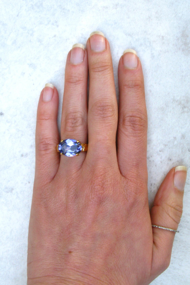 May include: A gold ring featuring a large, oval-cut, light purple gemstone set in a prong setting on the middle finger, accompanied by a thin, plain silver-toned band worn on the thumb, showcasing a contrast between the two distinct ring styles.