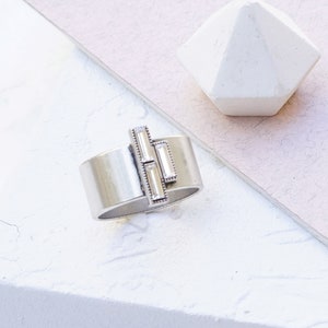 Puede incluir: Un anillo de plata con una banda ancha y tres piedras de cristal transparente engastadas en un patr&oacute;n geom&eacute;trico. CLEAR CRYSTAL | ANTIQUE SILVER