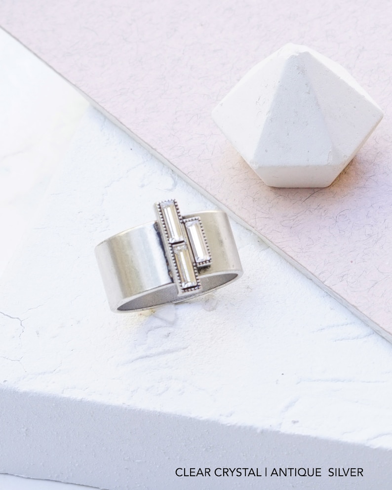 Puede incluir: Un anillo de plata con una banda ancha y tres piedras de cristal transparente engastadas en un patr&oacute;n geom&eacute;trico. CLEAR CRYSTAL | ANTIQUE SILVER