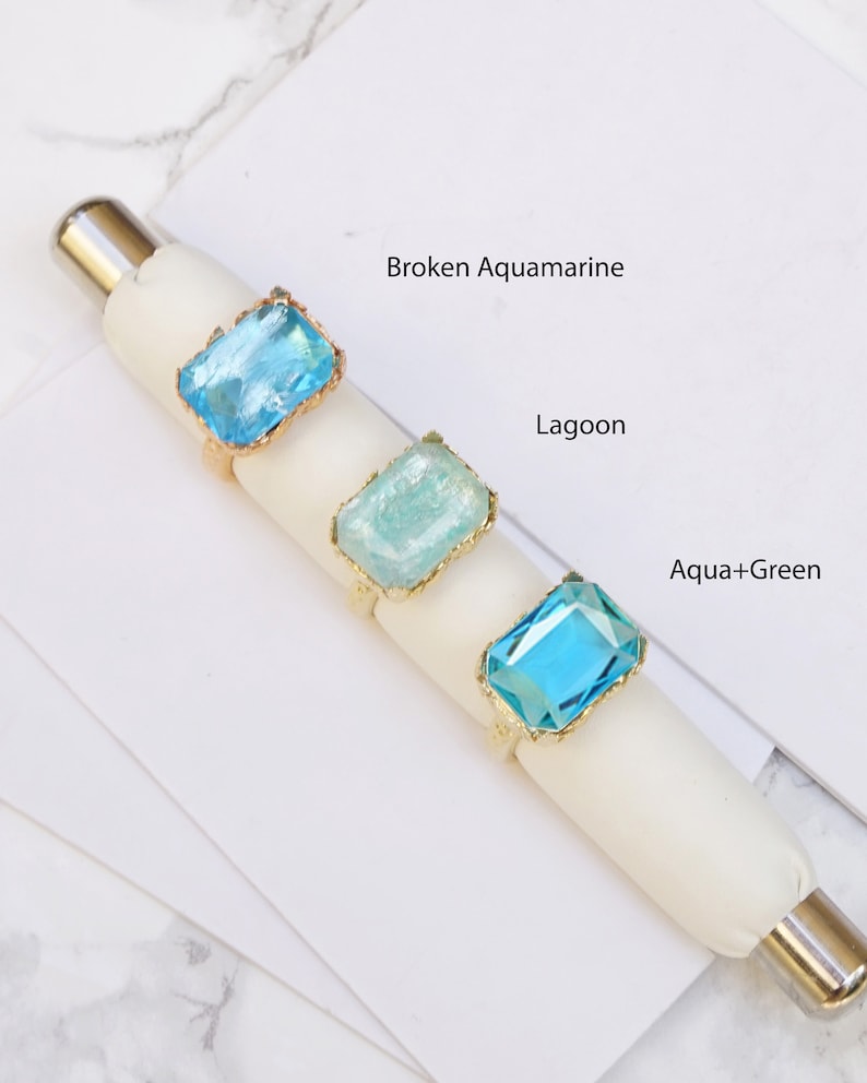 K&ouml;nnte beinhalten: Drei goldene Ringe mit verschiedenen Blaut&ouml;nen und gr&uuml;nen rechteckigen Steinen. Die Steine sind mit "Broken Aquamarine", "Lagoon" und "Aqua+Green" beschriftet.