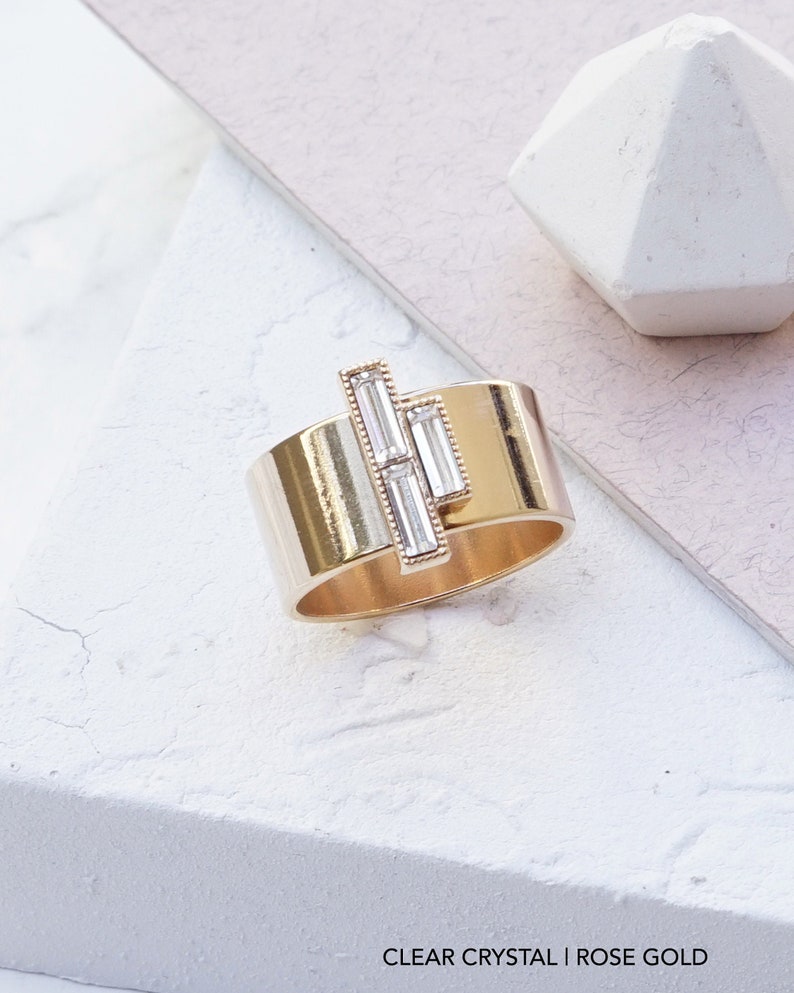 Puede incluir: Un anillo de oro rosa con un dise&ntilde;o de cristal transparente. El anillo es ancho y tiene una banda plana. El cristal est&aacute; engastado en una l&iacute;nea vertical y est&aacute; formado por tres piedras de forma baguette.