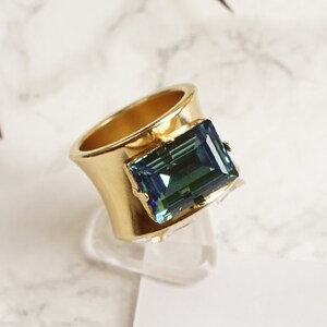 May include: A gold ring with a large, emerald green gemstone. The ring has a matte finish and the gemstone is set in a square shape.