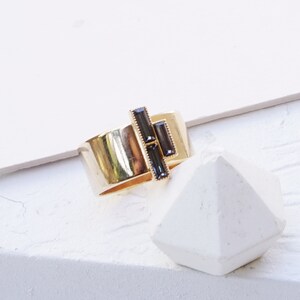 Puede incluir: Un anillo de oro rosa con un dise&ntilde;o geom&eacute;trico con tres piedras rectangulares negras. El anillo est&aacute; sobre una superficie blanca. SILVER NIGHT | ROSE GOLD