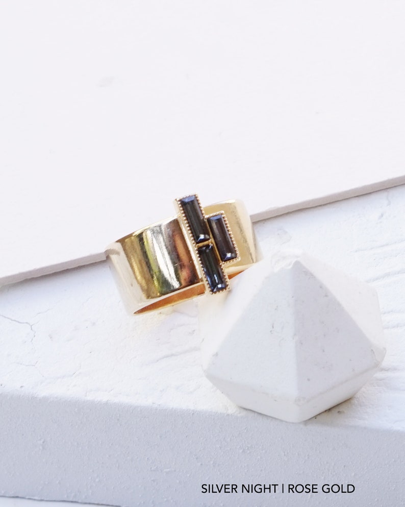 Puede incluir: Un anillo de oro rosa con un dise&ntilde;o geom&eacute;trico con tres piedras rectangulares negras. El anillo est&aacute; sobre una superficie blanca. SILVER NIGHT | ROSE GOLD