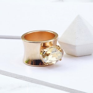 May include: A wide band ring featuring a rose gold finish and a golden shade. The ring is adorned with a single, large, oval-cut faceted stone set in a prong setting on the front. The text at the bottom reads: Color Golden shade with rose gold finish.