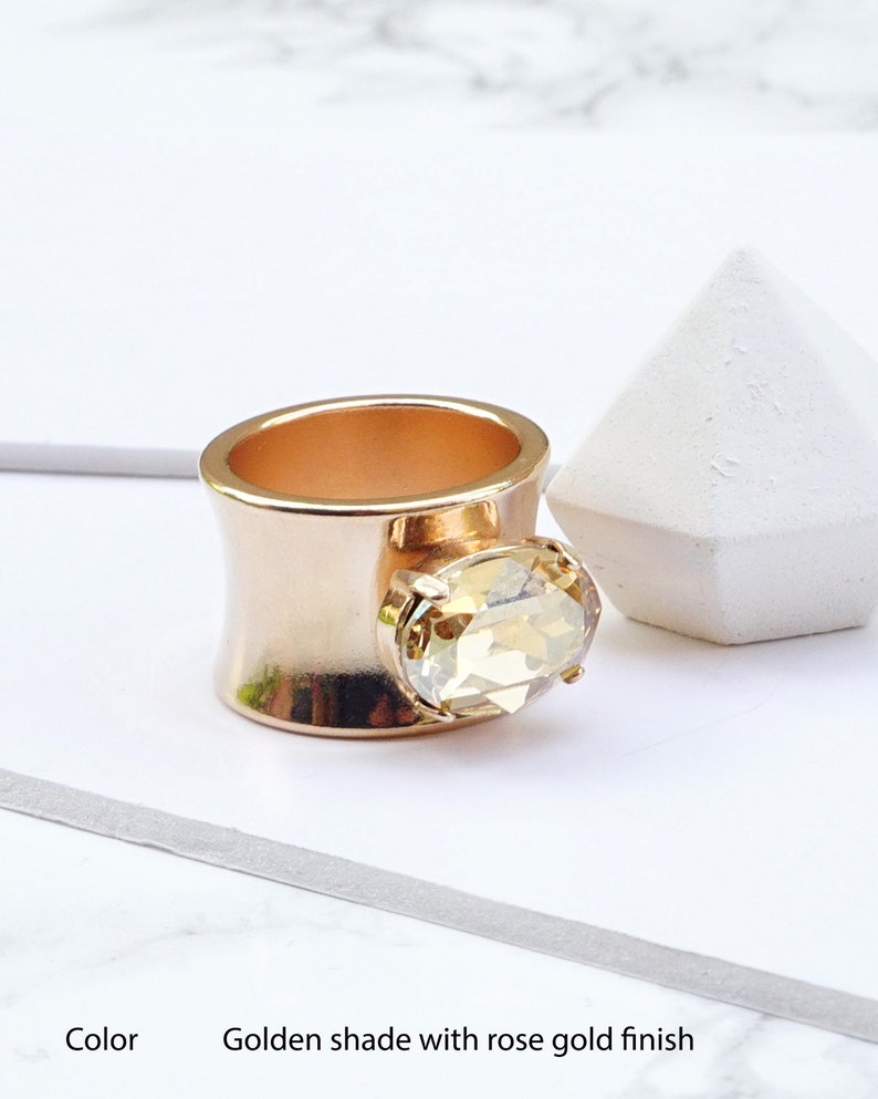 May include: A wide band ring featuring a rose gold finish and a golden shade. The ring is adorned with a single, large, oval-cut faceted stone set in a prong setting on the front. The text at the bottom reads: Color Golden shade with rose gold finish.