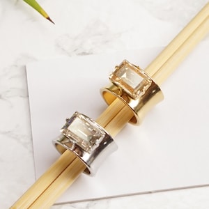 May include: Two wide band rings featuring rectangular emerald-cut crystals. One ring has a polished silver-tone finish with a clear crystal, and the other has a polished gold-tone finish with a light amber crystal. Text reads SILVER SHADE SHAINY SILVER and GOLDEN SHADOW SHAINY GOLD.