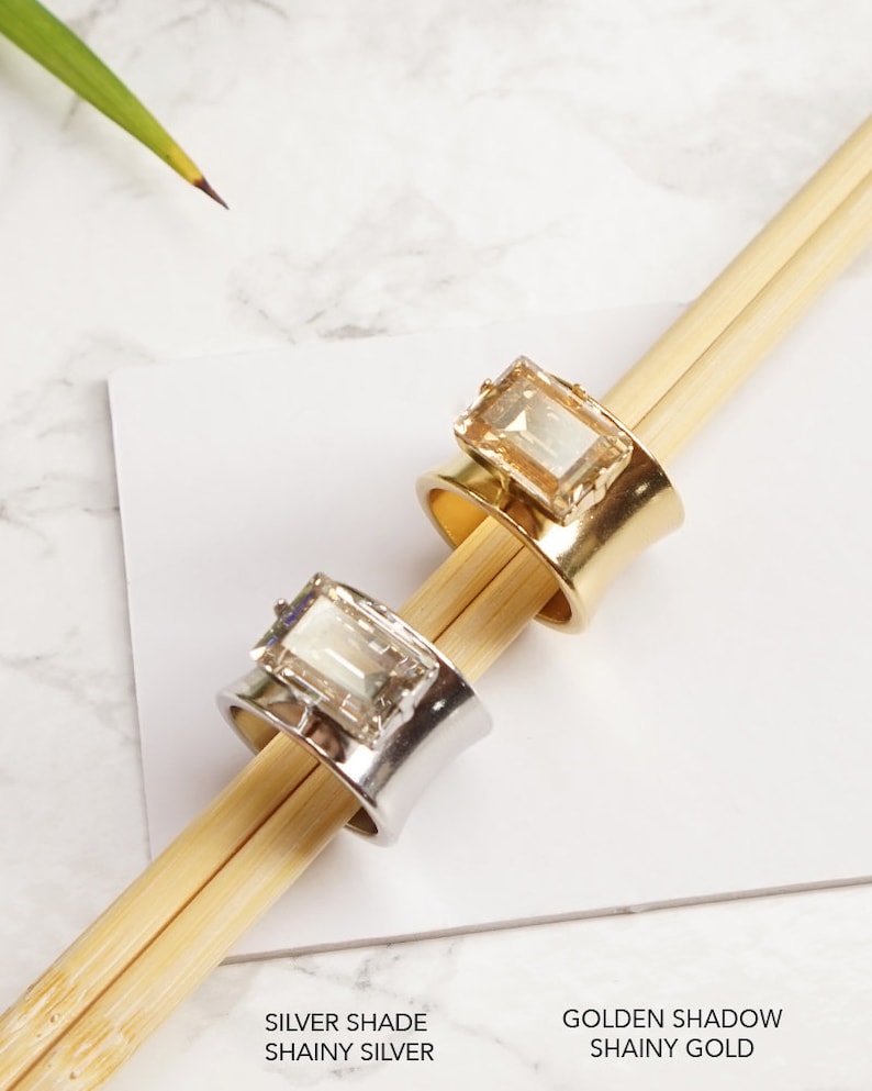 May include: Two wide band rings featuring rectangular emerald-cut crystals. One ring has a polished silver-tone finish with a clear crystal, and the other has a polished gold-tone finish with a light amber crystal. Text reads SILVER SHADE SHAINY SILVER and GOLDEN SHADOW SHAINY GOLD.