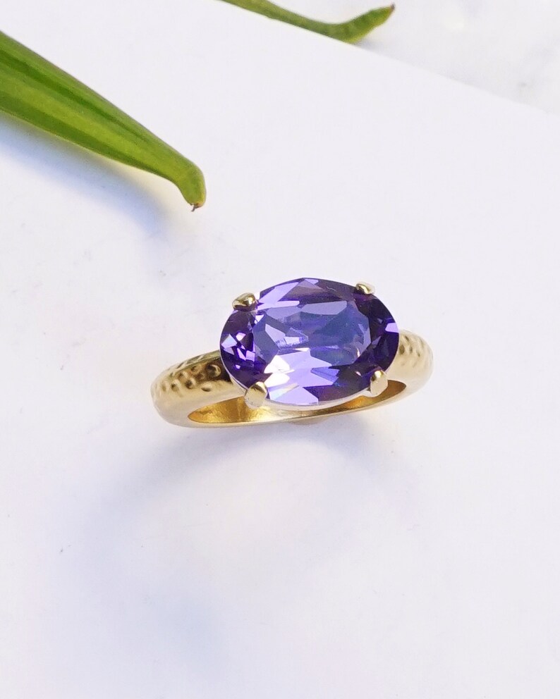 May include: A gold ring featuring a large, oval-cut purple gemstone set in four prongs. The band of the ring has a textured, hammered finish, providing a distinct contrast to the smooth, faceted surface of the vibrant purple stone.