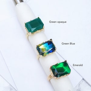 May include: Three gold rings with different colored stones. The top ring has a green opaque stone, the middle ring has a green and blue stone, and the bottom ring has a dark green stone. The rings are labeled "Green opaque", "Green Blue", and "Emerald".