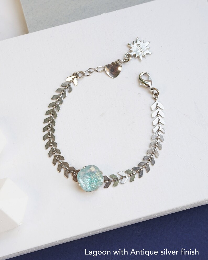 Puede incluir: Una pulsera de tono plateado con una cadena de eslabones en forma de chevr&oacute;n y una piedra cuadrada de color azul claro texturizada. La pulsera incluye un cierre de mosquet&oacute;n, un dije de coraz&oacute;n y un dije de estrella con el texto Lagoon.