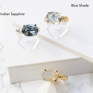 May include: Three rings with oval-shaped stones and textured bands. One ring features an Indian Sapphire stone on a silver-toned band, another has a Blue Shade stone on a gold-toned band, and the third has a Sand Opal stone on a gold-toned band.