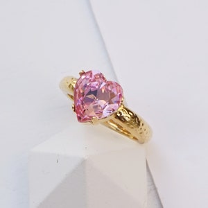 May include: A gold-toned ring featuring a faceted, heart-shaped light pink gemstone set in a prong setting. The band has a textured, hammered finish. The text "Light rose with Shiny gold finish" is printed at the bottom of the image.