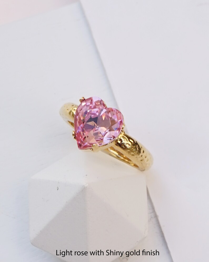 May include: A gold-toned ring featuring a faceted, heart-shaped light pink gemstone set in a prong setting. The band has a textured, hammered finish. The text "Light rose with Shiny gold finish" is printed at the bottom of the image.