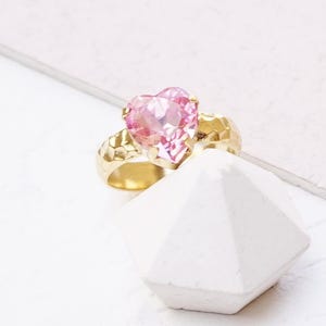 May include: A ring featuring a faceted heart-shaped light rose gemstone set in a hammered matte gold band. The text LIGHT ROSE | MATTE GOLD is printed at the bottom of the image. The ring is positioned on a white geometric surface.
