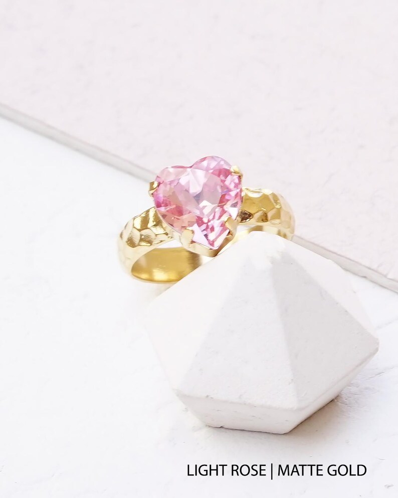 May include: A ring featuring a faceted heart-shaped light rose gemstone set in a hammered matte gold band. The text LIGHT ROSE | MATTE GOLD is printed at the bottom of the image. The ring is positioned on a white geometric surface.