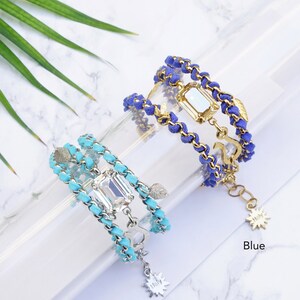Op de afbeelding: Twee gelaagde armbanden met een gouden ketting en turquoise of blauw su&egrave;de koord, elk met een heldere gefacetteerde edelsteen en een gouden bedel. De turquoise armband heeft een sterbedel en de blauwe armband heeft een ankerbedel. De armbanden worden getoond op een wit marmeren oppervlak.