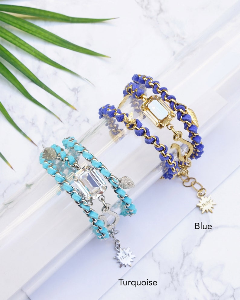 Op de afbeelding: Twee gelaagde armbanden met een gouden ketting en turquoise of blauw su&egrave;de koord, elk met een heldere gefacetteerde edelsteen en een gouden bedel. De turquoise armband heeft een sterbedel en de blauwe armband heeft een ankerbedel. De armbanden worden getoond op een wit marmeren oppervlak.