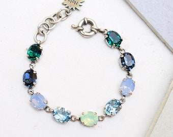 May include: A silver bracelet with a charm that says "Holy" and features a variety of oval-shaped gemstones in shades of blue, green, and white. The bracelet is made with a chain link design and a lobster clasp.