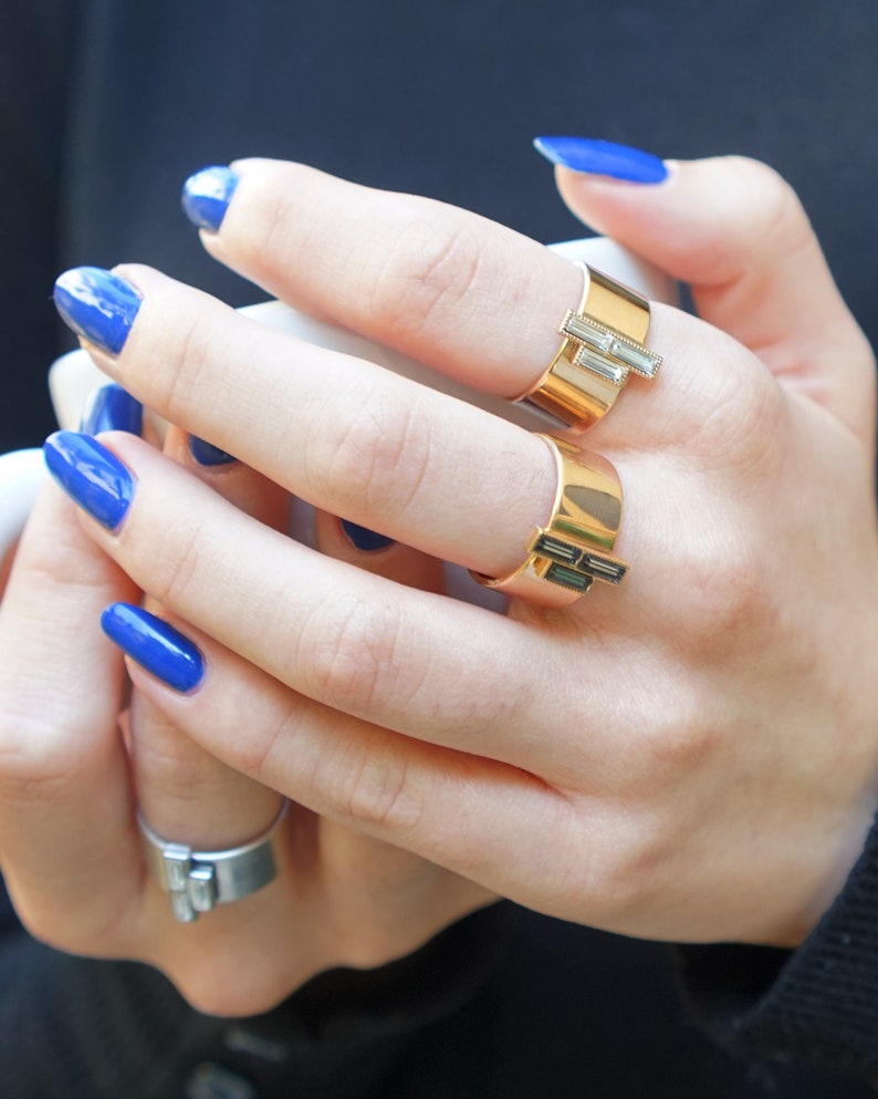 Puede incluir: Dos anillos de oro con piedras rectangulares en una mano con esmalte de u&ntilde;as azul.
