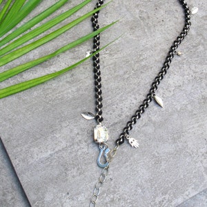 Op de afbeelding: Een zwarte ketting met zilveren bedels, waaronder bladeren, een hand en een grote, heldere kristal. De ketting heeft een zilveren sluiting en een zilveren sterbedel die aan een ketting hangt.