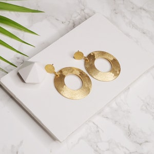 May include: A pair of matte gold hoop earrings with a textured finish. The earrings are large and circular, with a slightly irregular shape.