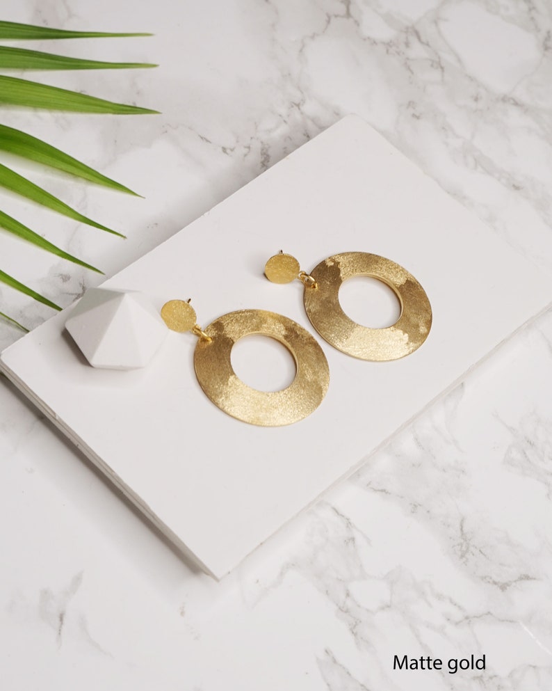 May include: A pair of matte gold hoop earrings with a textured finish. The earrings are large and circular, with a slightly irregular shape.