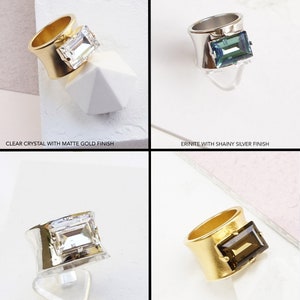 May include: A collection of four rings featuring rectangular crystals in clear, erinite, and smokey quartz colors. The bands are finished in matte gold, shiny silver, or shiny gold. Text labels include Clear Crystal with Matte Gold Finish, Erinite with Shainy Silver Finish, Clear Crystal | Shiny Silver, and Smokey Quartz | Matte Gold.