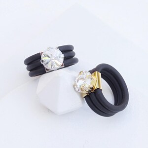 May include: Two black silicone rings with a clear crystal stone. The ring on the left has a silver setting, and the ring on the right has a gold setting. CLEAR SILVER | CLEAR YELLOW GOLD