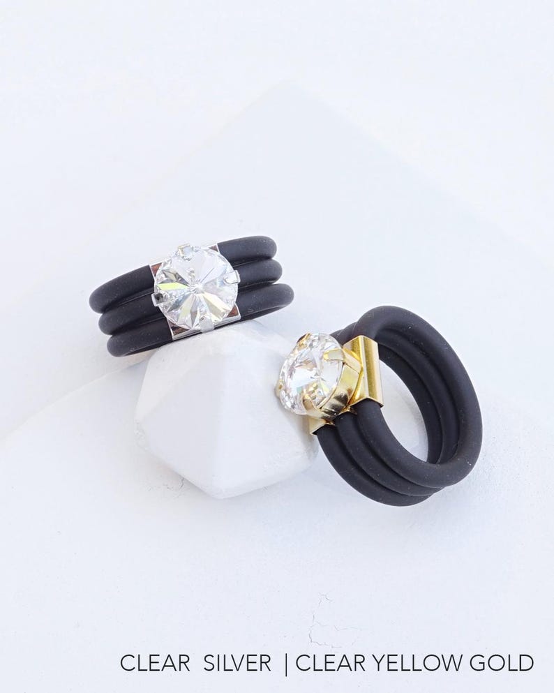 May include: Two black silicone rings with a clear crystal stone. The ring on the left has a silver setting, and the ring on the right has a gold setting. CLEAR SILVER | CLEAR YELLOW GOLD