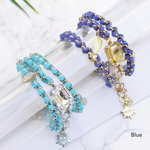 Op de afbeelding: Twee gelaagde armbanden met een gouden ketting en su&egrave;de koord. De ene armband is turquoise met een helder kristal en de andere is blauw met een gouden kristal. Beide armbanden hebben een gouden bladeren bedel en een gouden ster bedel met het woord "Holy" erop.