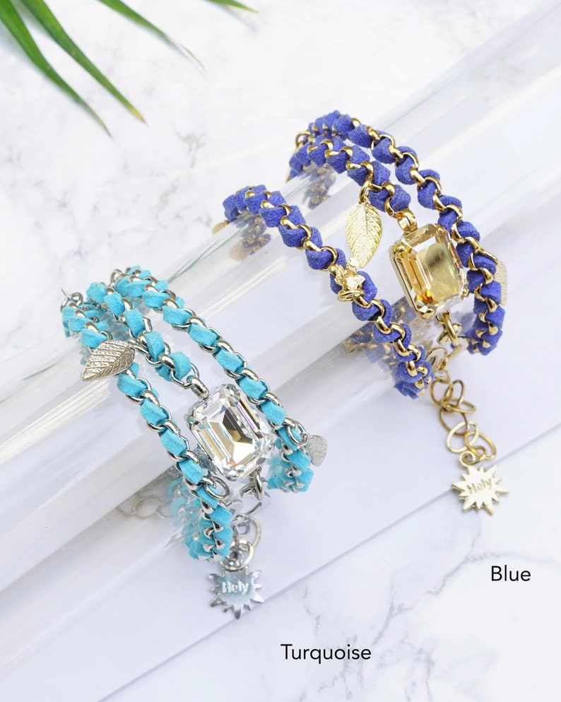 Op de afbeelding: Twee gelaagde armbanden met een gouden ketting en su&egrave;de koord. De ene armband is turquoise met een helder kristal en de andere is blauw met een gouden kristal. Beide armbanden hebben een gouden bladeren bedel en een gouden ster bedel met het woord "Holy" erop.