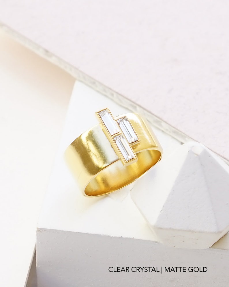 Puede incluir: Un anillo de oro con acabado mate y tres cristales transparentes en forma de baguette engastados en la banda. CLEAR CRYSTAL | MATTE GOLD